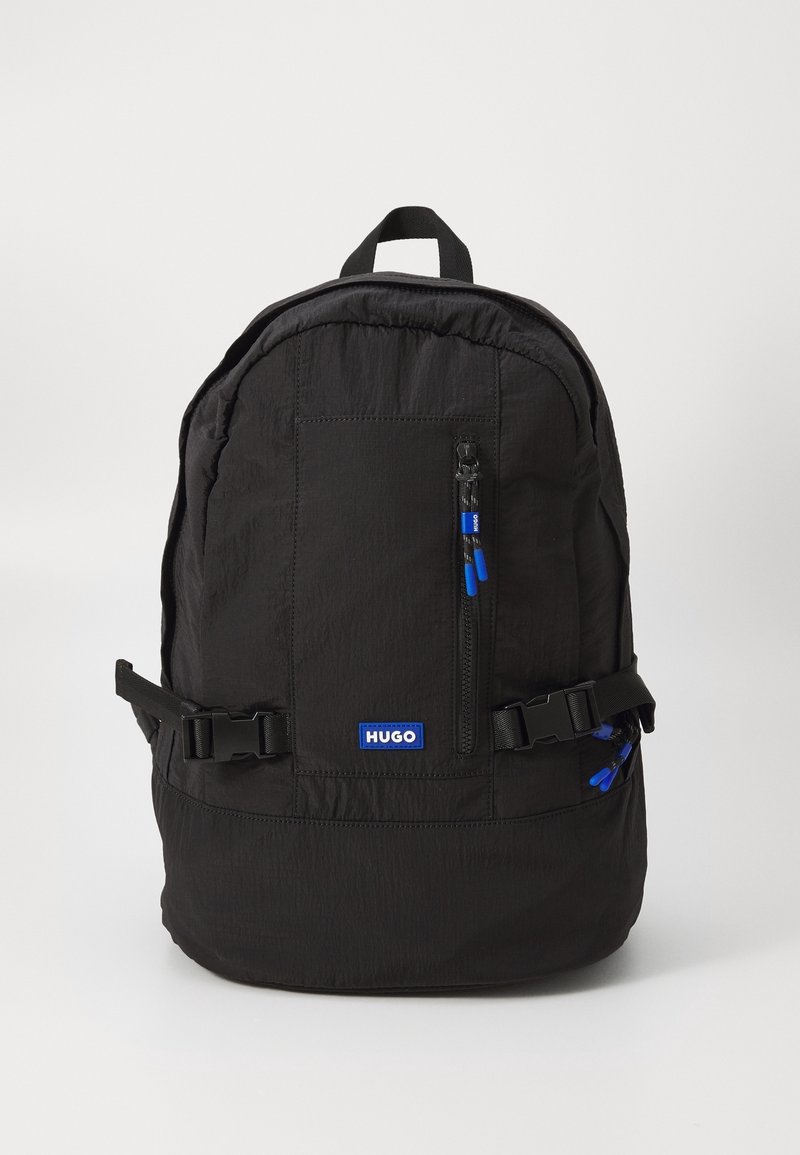 Sac à dos noir Hugo avec poche avant zippée, bretelles latérales ajustables et tirettes de fermeture éclair bleues sur fond blanc uni.