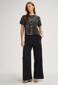 Svart kortärmad topp med ett guldfärgat mönster, kombinerad med löst sittande svarta byxor med rött logo och fickor. Platta sandaler.