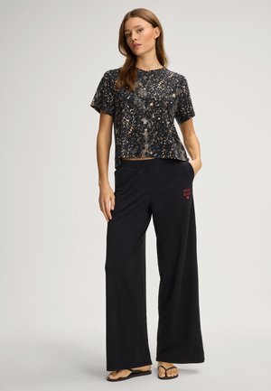 Zwart, kortärmelig top met een goudkleurig patroon, gecombineerd met loszittende zwarte broek met een rood logo en zakken. Platte sandalen.