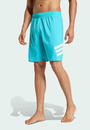 Muž oblečený v tyrkysových plavkových šortkách s tromi bielymi diagonálnymi pruhmi na ľavej nohe, stojaci bosý proti jednoliatej stene.