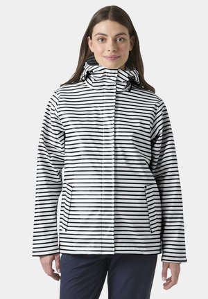 Mujer con chaqueta blanca de rayas horizontales negras y capucha, de pie y mirando hacia adelante sobre un fondo liso.