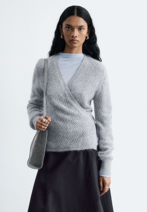 Mujer joven con cabello largo y negro, que lleva un suéter envolvente texturizado de color gris claro, falda negra y sostiene un bolso gris de hombro.