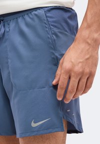 Gros plan sur la main d'une personne reposant près de la poche latérale d'un short de sport bleu Nike avec un logo visible.