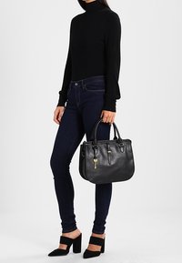 Sac à main en cuir noir au design structuré, avec des poignées supérieures et des accents en métal doré. Modèle portant un col roulé noir et un jean foncé.