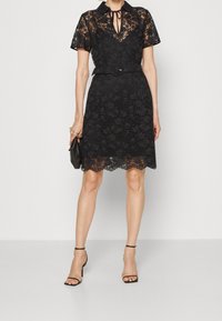 Robe noire en dentelle avec col, manches courtes et taille ceinturée. Motif floral et détail d'ourlet festonné. Paire de chaussures à talons noires à lanières.