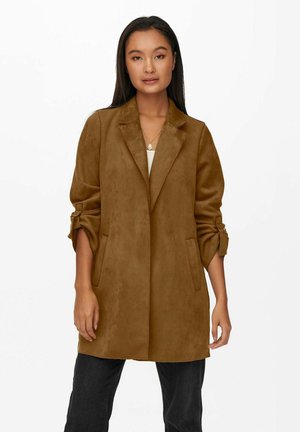 Femme portant un long blazer en daim marron avec des manches retroussées, un haut blanc, un pantalon noir et un collier pendentif sur fond blanc.