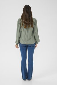 Blusa verde fluida con maniche a sbuffo e polsini ricamati, abbinata a jeans blu aderenti che si allargano sul fondo.