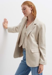 Blazer beige in lino con una vestibilità leggermente oversize, design monopetto, rever a punta e due tasche frontali. Indossato sopra un gilet.
