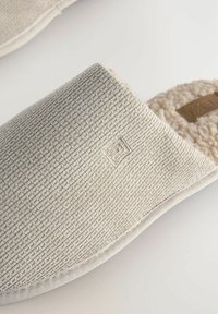 Graue Stoffschlappen mit einer strukturierten Oberfläche, weichem Innenfutter und gesteppten Kanten. Verfügt über ein kleines, geprägtes Logo an der Seite.