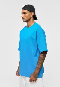 Felle blauwe t-shirt met korte mouwen van katoen, met een ronde hals en een relaxte pasvorm. Draagt een zwarte pet en toont arm tattoo's.