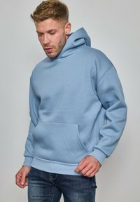 Jeune homme regardant vers le bas, portant un sweatshirt bleu clair et un jean sombre, sur un fond gris uni.