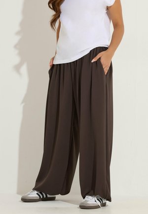 Mujer con pantalones anchos de pierna ancha sueltos de color marrón oscuro, camiseta blanca y zapatillas blancas con rayas negras, de pie con las manos en los bolsillos.