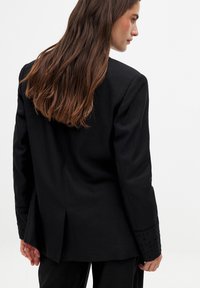 Schwarzer Blazer mit taillierter Passform, der detailliert bestickte Manschetten und einen Schlitz auf der Rückseite verfügt. Glatte Textur und strukturiertes Design.