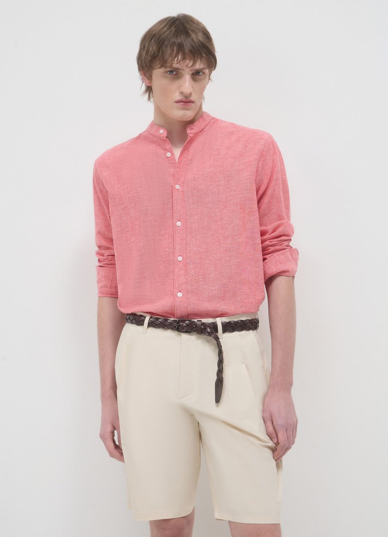 Chemise en lin rose avec col mandarin et fermeture boutonnée, associée à un short crème et une ceinture tressée marron. Texture lisse et coupe décontractée.
