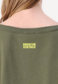 Olívazöld pulóver széles nyakkivágással, bordázott szegéllyel és sárga "AMERICAN VINTAGE" logóval a háton. Puha, sima tapintás.