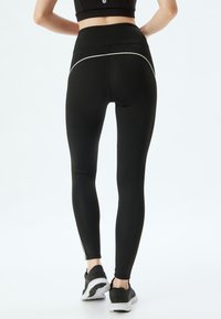 Zwarte high-waisted leggings met een gladde textuur, voorzien van witte zijaccenten en een strakke pasvorm, gecombineerd met zwarte sportschoenen.