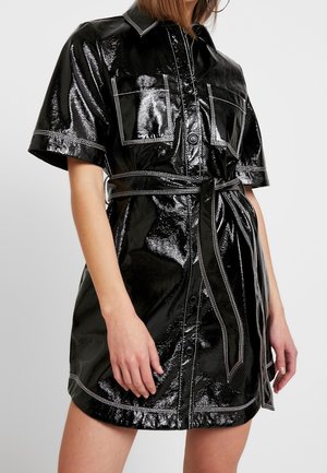 Robe-chemise en similicuir noir brillant, manches courtes, deux poches poitrine, fermeture à boutons et taille ceinturée. Détails de surpiqûres blanches.