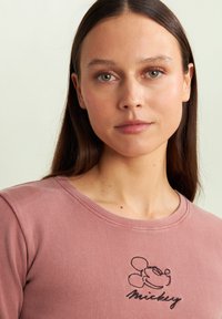 Maglietta a maniche lunghe rosa chiaro a coste con scollo rotondo e motivo ricamato di Topolino nero sul petto. Texture liscia.