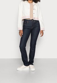 Mujer vestida con pantalones ajustados de mezclilla azul oscuro, chaqueta corta blanca con rayas rosas, top beige y zapatillas blancas, de pie sobre un suelo gris.