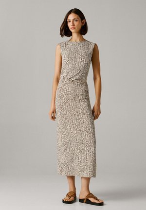 Frau in ärmellosem, beige gemustertem Midikleid mit Details an der Taille und braunen flachen Sandalen, stehend vor einem einfarbigen Hintergrund.