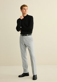 Schwarzes Langarmhemd, hellgraue Hose mit strukturiertem Finish, schwarzer Gürtel und schwarze Anzugschuhe. Modell steht vor einem neutralen Hintergrund.