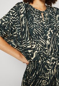 Femme portant un chemisier ample imprimé zèbre noir et blanc et un pantalon assorti avec taille froncée, debout avec une main sur la hanche.