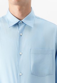 Nahaufnahme eines hellblauen Hemds mit Kragen, das weiße Knöpfe und eine Brusttasche aufweist, vor einem neutralen Hintergrund.