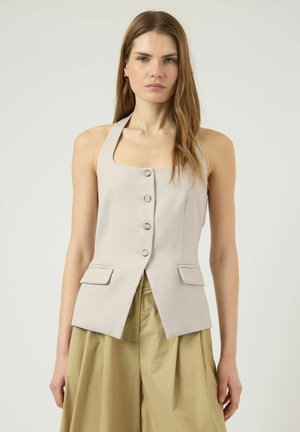 Kobieta z długimi włosami, ubrana w bezrękawnik w beżowym kolorze zapinany na guziki oraz luźne, szerokie spodnie khaki z wysokim stanem, stojąca na tle jednolitego, jasnego tła.