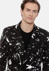 Młody mężczyzna o jasnej skórze i jasnobrązowych włosach, ubrany w czarną marynarkę z białymi abstrakcyjnymi plamami farby na czarnej koszuli, patrzy w aparat.