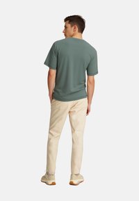 Rövid ujjú, kerek nyakú póló finom zöld színben, világos bézs nadrággal és bézs sportcipővel párosítva, hátulról látható.