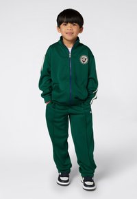 Niño pequeño de pie con las manos en los bolsillos, vistiendo un chándal verde con chaqueta de cremallera y zapatillas negras y blancas sobre un fondo liso.