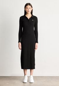 Μαύρο μακρύ μανίκι πόλο φόρεμα με εφαρμοστή σιλουέτα, ραβδωτή υφή και κολάρο με κουμπιά. Διαθέτει μικρό πράσινο λογότυπο στο στήθος.