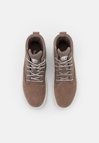 Een paar bruine suède wandelschoenen met witte gemusterde veters en witte zolen, vanuit bovenaf bekeken, op een witte achtergrond.