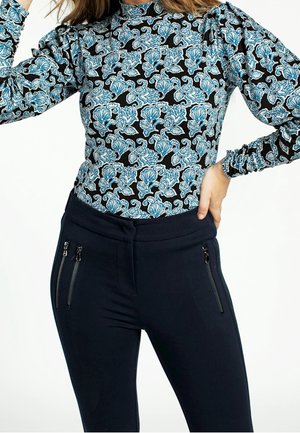 Femme portant un haut noir ajusté avec un motif floral bleu et blanc, et un pantalon noir taille haute avec des poches à fermeture éclair verticales.