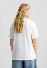 T-shirt bianco in cotone con maniche corte, vestibilità rilassata, scollatura rotonda e orlo dritto, mostrato da dietro con capelli biondi mossi visibili.