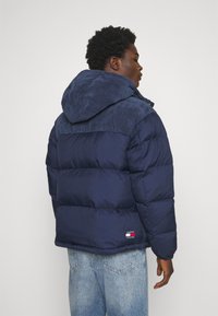 Giacca puffer blu navy con cappuccio in corduroy, texture trapuntata e polsini elasticizzati, con una piccola etichetta logo sui lati.