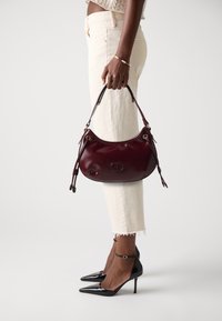 Burgundy leather handbag with a curved shape, short handles, and decorative drawstrings; worn with white cropped pants and black stiletto heels.