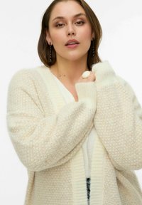 Adīts kardigans krēmkrāsā ar teksturētu rakstu, platām piedurknēm, V veida izgriezumu un ribotu apdari uz aprocēm un apakšmalu.