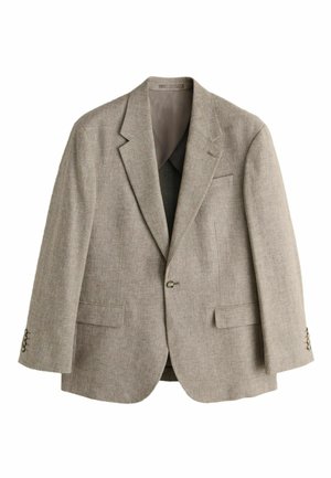 Ung mand med kort krøllet hår iført en beige dragt og en mørkegrøn sweater, der står mod en ensfarvet beige væg.