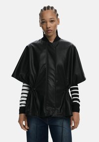 Veste en cuir synthétique noir avec un design ample et capé, présentant des cordons de serrage sur les côtés et un col noir à côtes, portée sur une chemise à rayures à manches longues.