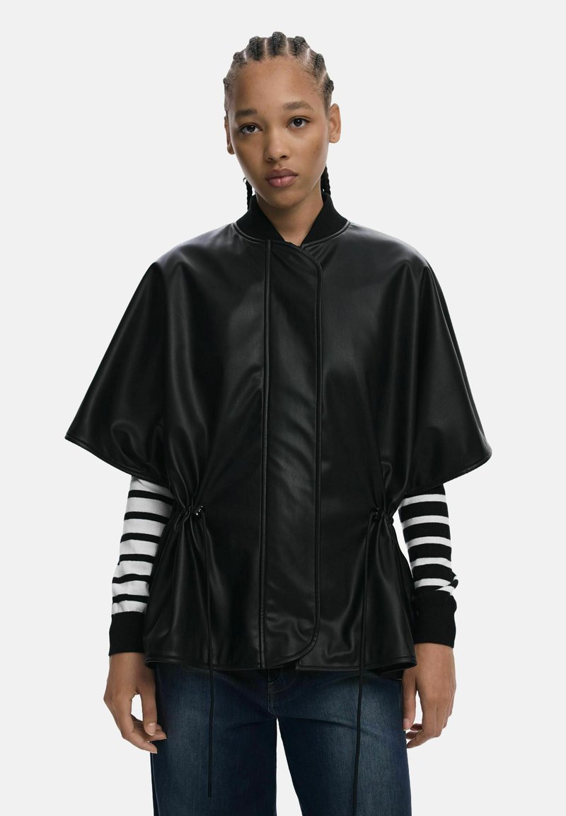 Veste en cuir synthétique noir avec un design ample et capé, présentant des cordons de serrage sur les côtés et un col noir à côtes, portée sur une chemise à rayures à manches longues.
