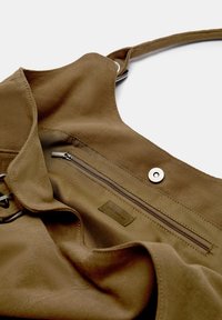 Interno di una borsa a tracolla in tessuto marrone, dotato di una tasca con zip e un'etichetta. Il materiale principale ha un aspetto testurizzato e resistente.