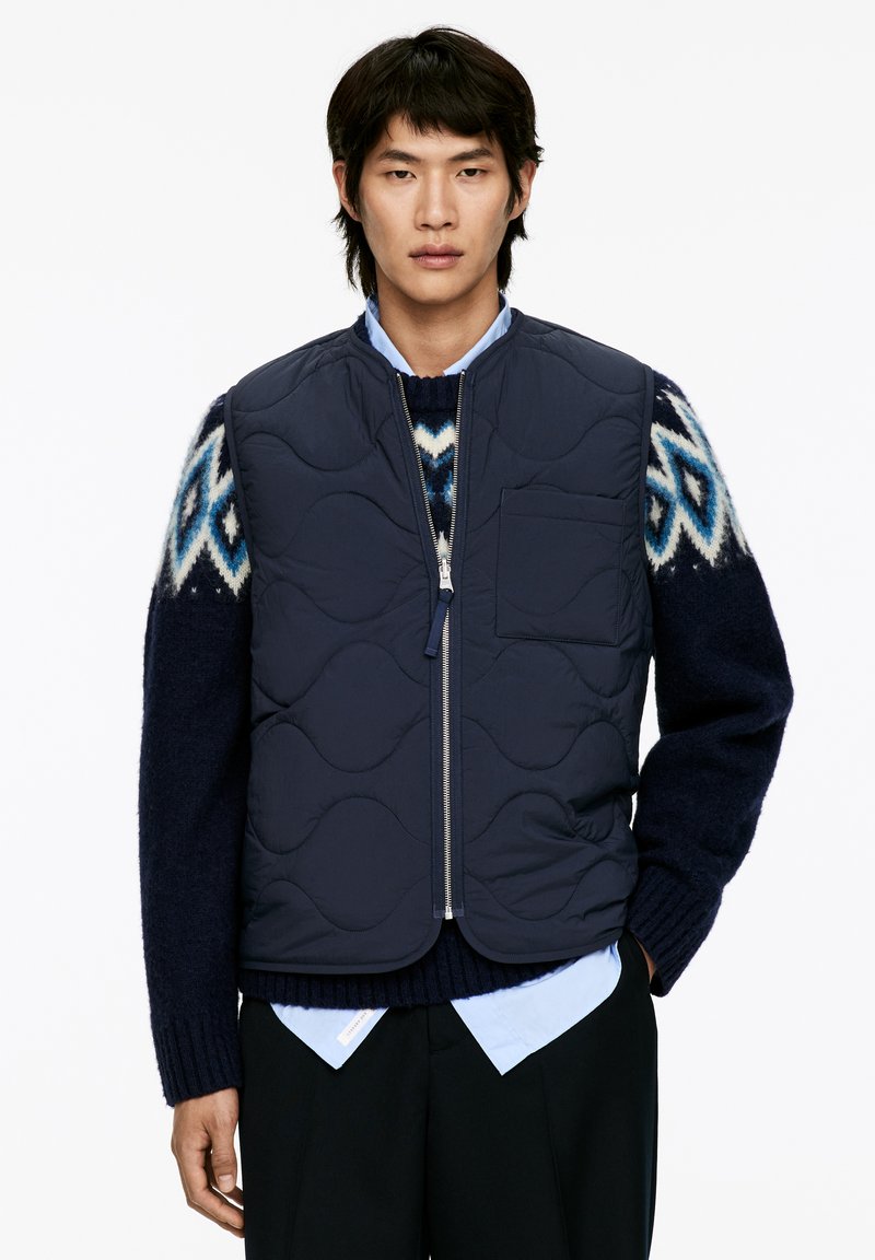 Gilet matelassé marine avec un col haut, une poche frontale et une fermeture éclair argentée, porté par-dessus un pull à motifs et une chemise bleu à col.