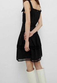 Abito nero a strati con spalline, bordi in pizzo e dettagli intricati con ritagli all'orlo. Il modello indossa stivali bianchi fino al ginocchio.