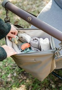 Grå pusletaske med en lynlåslomme indeholder bløde tæpper, en sut og legetøj, hvilende på en klapvogns håndtag i en naturlig omgivelser.