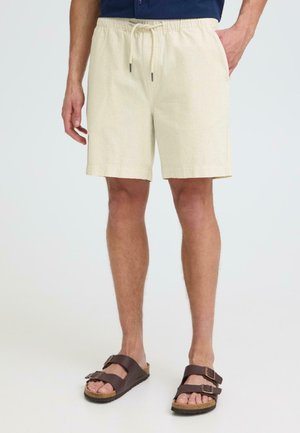 Homme portant un short à cordon de serrage couleur crème et des sandales marron foncé à double bride, debout sur un fond blanc.