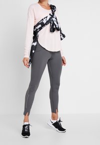 Lichtroze lange mouwen top, grijze legging met zijslits, zwarte gebandeerde sjaal geknoopt om de taille, en zwarte sportieve schoenen met witte accenten.