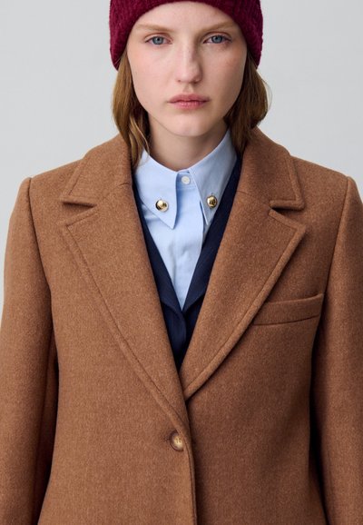 Cappotto in misto lana marrone con rever a punta, chiusura a singolo bottone e taschino sul lato sinistro del petto; indossato sopra una camicia azzurro chiaro e un gilet scuro.