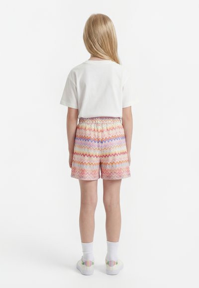 Niño con cabello largo y rubio de espaldas, vestido con una camiseta blanca, pantalones cortos de colores con patrón de zigzag, calcetines blancos y zapatillas blancas.
