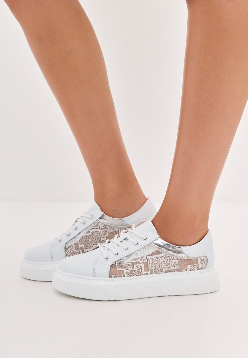 Sneakers bianchi con un pannello laterale beige a fantasia, dettagli argentati e texture su una suola bianca spessa. Design con lacci, struttura bassa.